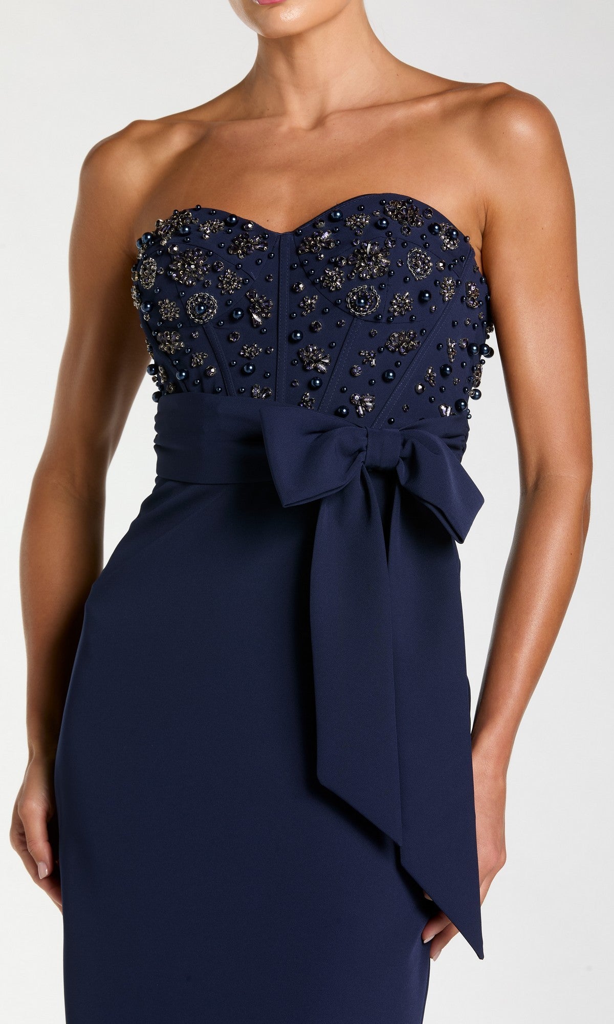 A woman wearing the Mac Duggal 2395 long formal dress, a strapless gown in navy blue adorned with bead embellishments and a large bow at the waist.